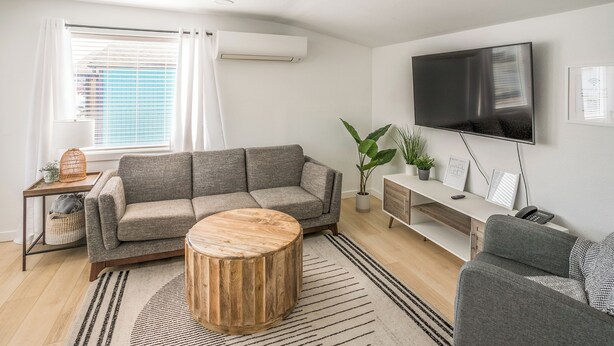 A modern living room with a grey sofa, a wooden coffee table, a TV mounted on the wall, and a small side table with a plant.