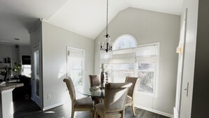 Un coin repas avec une table en verre, quatre chaises et un lustre. De grandes fenêtres sont ornées de rideaux blancs, et la pièce est dotée d’un plafond haut.