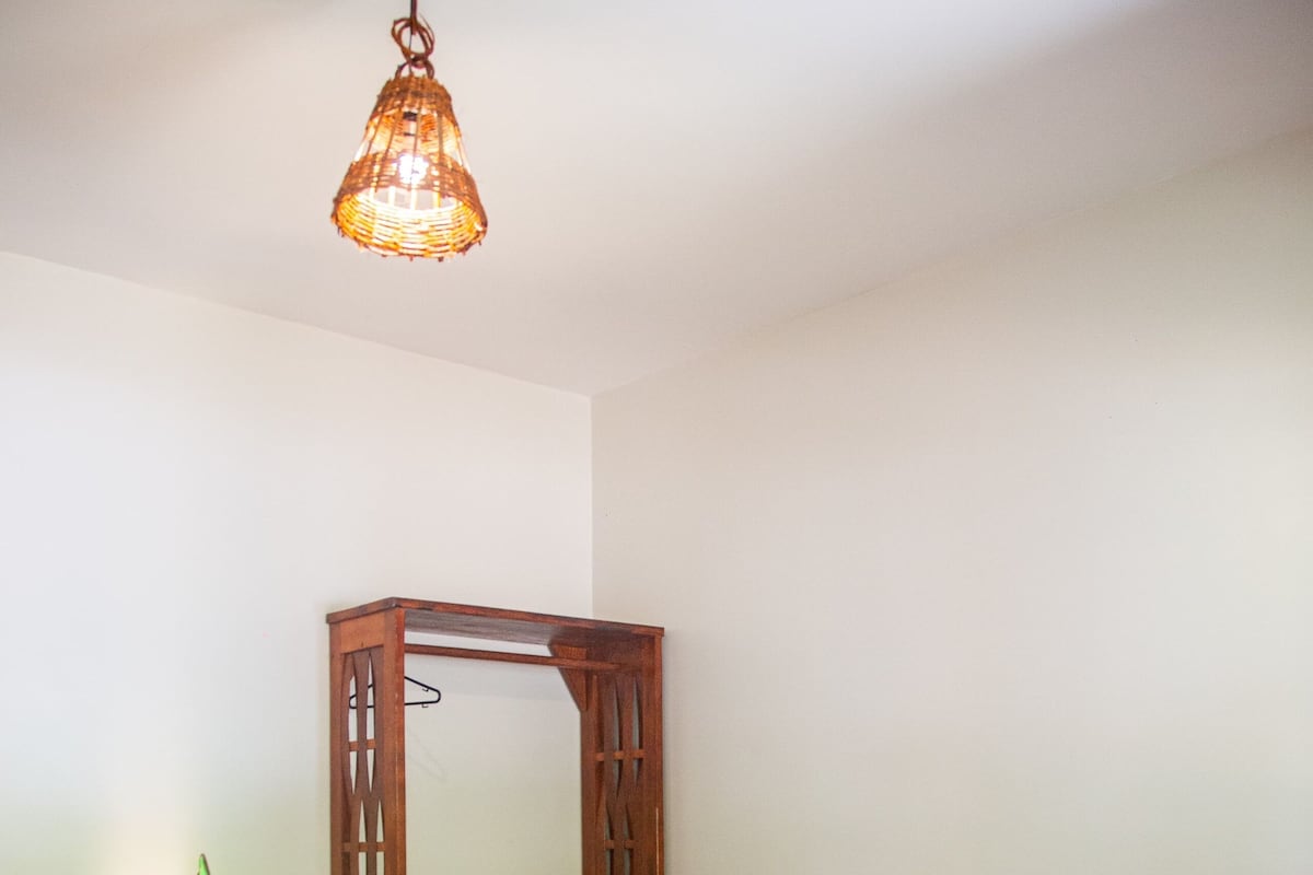 Una cama bien hecha con un edredón a cuadros, un espejo de madera colgante y una lámpara de pie.
