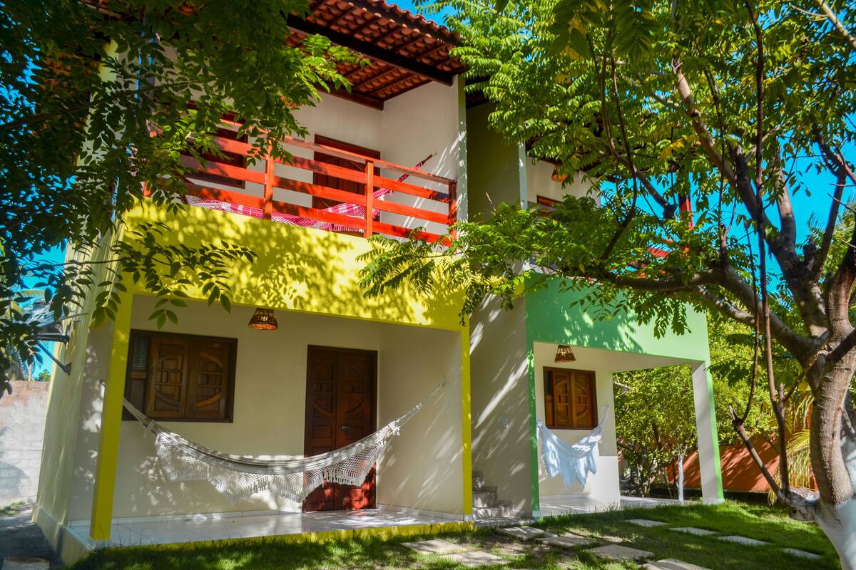 Una casa de dos pisos con una hamaca en el porche, un balcón con barandas rojas y una puerta verde.