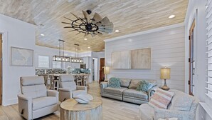 A spacious living room with a modern ceiling fan, a dining area with a kitchen in the background, and a large window providing natural light.