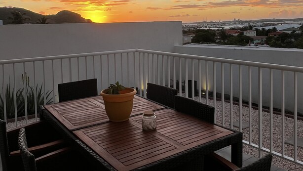 A balcony with a wooden table and chairs, a potted plant, and a view of a sunset over a cityscape.