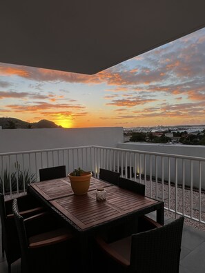 A balcony with a wooden table and chairs, a potted plant, and a view of a sunset over a cityscape. - Adorable Private Room with partial Sea View (Sentry Hill)