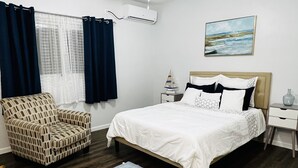 A bedroom with a bed, a chair, a nightstand, a wall-mounted air conditioner, a window with curtains, and a painting.