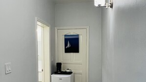 A narrow hallway with a white door at the end, a small refrigerator, and a wall-mounted light fixture.