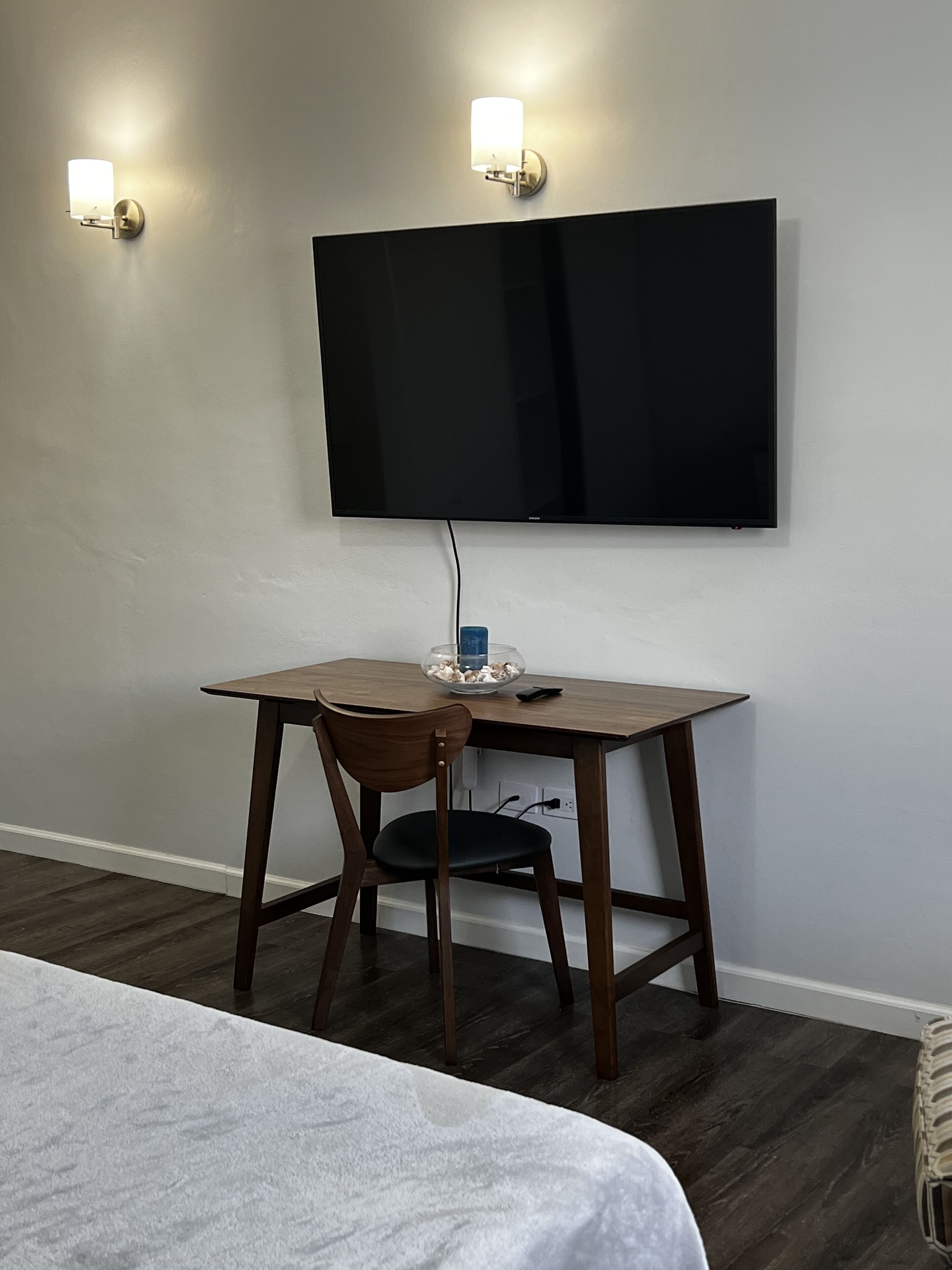 A room with a bed, a desk with a chair, a flat-screen TV mounted on the wall, and two wall-mounted lights.
