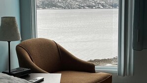 Un dormitorio con vista a un lago y colinas, un sillón marrón, una lámpara y una mesita de noche con un control remoto.
