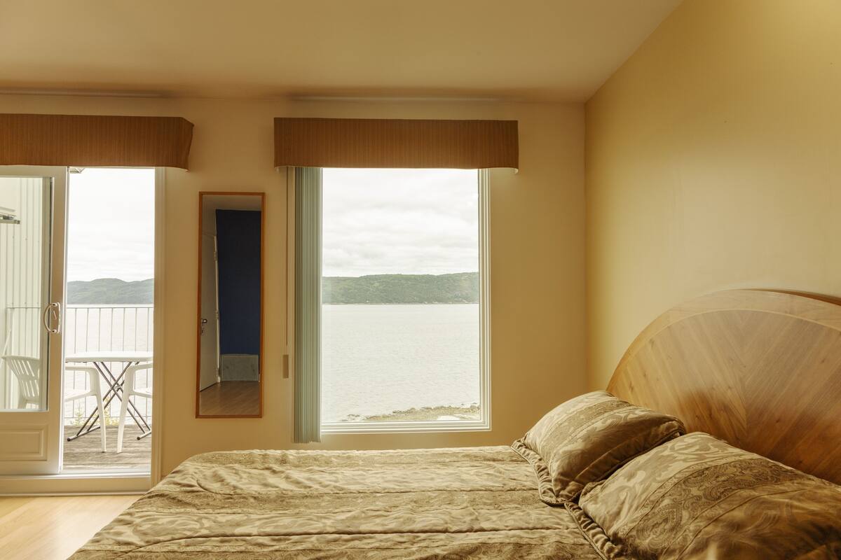 Ein Schlafzimmer mit Bett, Fenster mit Blick auf ein Gewässer und einer Tür zu einem Außenbereich mit Tisch und Stühlen.
