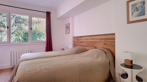 A bedroom with two beds, a window with a view of greenery, a radiator, and a bedside table with a lamp.