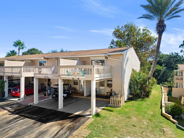 A two-story building with a covered parking area, a balcony, and a palm tree in the background. - Spacious 2-Level End Unit Just Steps From the Beach With Private Patio & Balcony (Panama City Beach)