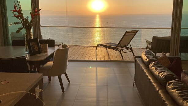 Ein Balkon mit Blick auf den Sonnenuntergang über dem Meer, ein Essbereich mit Tisch und Stühlen sowie eine Couch.