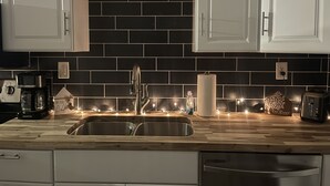 Een keuken met witte kastjes, een zwart betegelde achterwand, een roestvrijstalen spoelbak en houten aanrechtbladen.