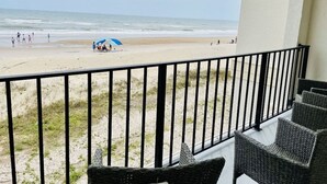Un balcon doté de meubles en osier, donnant sur une plage où se trouvent des gens et un parasol bleu.