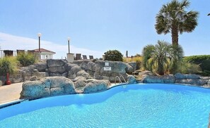 A swimming pool with a stone wall, palm trees, and a building with a red roof in the background. - New Listing "Sol Intention" 3 Bedroom Villa Capriani, fully renovated (North Topsail Beach)
