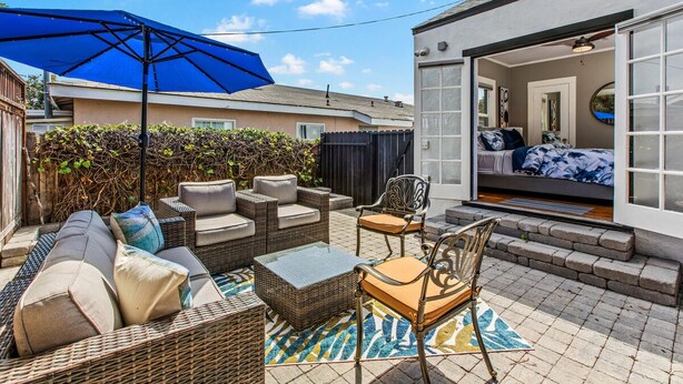 Eine Terrasse mit Korbmöbeln, einem blauen Sonnenschirm und Blick in ein Schlafzimmer mit Bett und Spiegel.