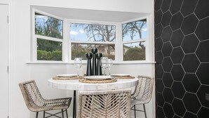 Un rincón comedor con una mesa blanca para cuatro personas, rodeado de sillas de mimbre y una pared de baldosas hexagonales negras.
