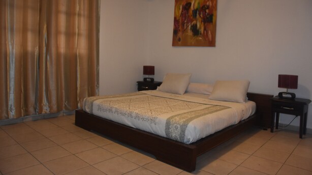 A bedroom with a large bed, two bedside tables, a painting on the wall, and a window with curtains. - FURNISHED HOUSE COSY (Libreville)