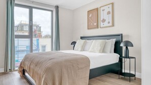 Un dormitori amb un llit, una tauleta de nit, una finestra amb cortines i dos quadres al mur.