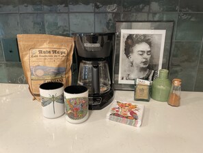 Uma bancada de cozinha com uma cafeteira, um pacote de café, duas canecas, uma fotografia emoldurada e um objeto decorativo pequeno.