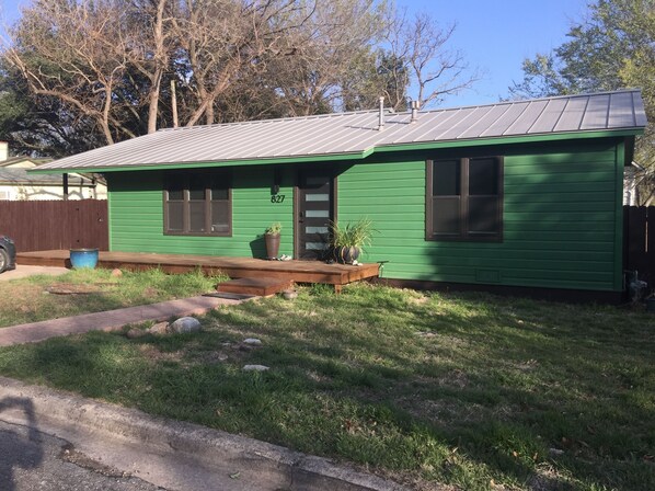 Uma casa térrea verde com telhado de metal, varanda frontal e o número 827 na porta.