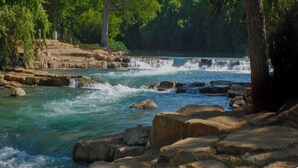 A river with a waterfall, surrounded by rocks and trees. - Walk to the crystal clear San Marcos river (San Marcos)