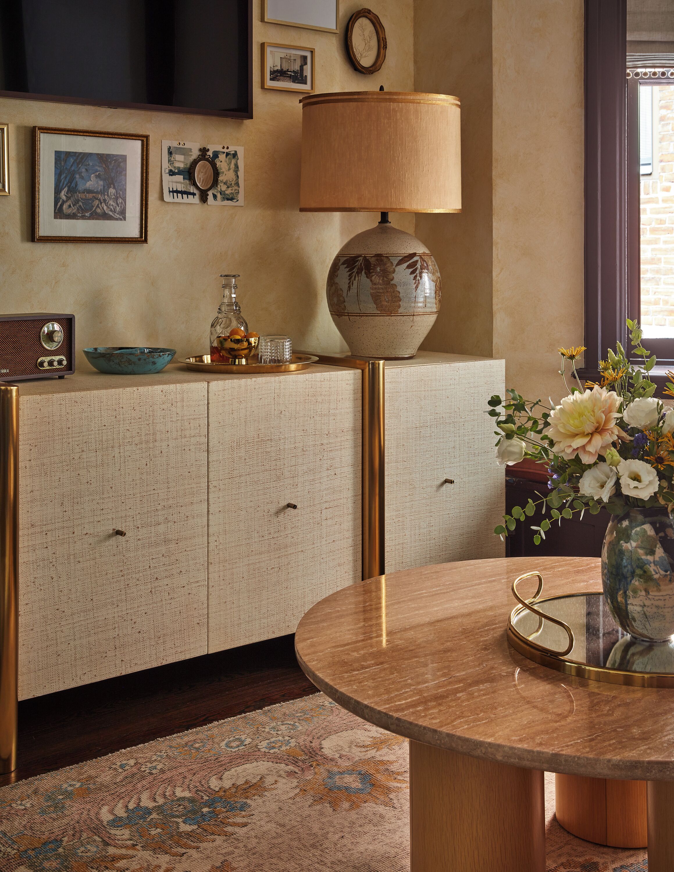 Une pièce avec une table ronde en bois, un vase rempli de fleurs, une radio ancienne et une lampe décorative posée sur un meuble à la texture particulière.