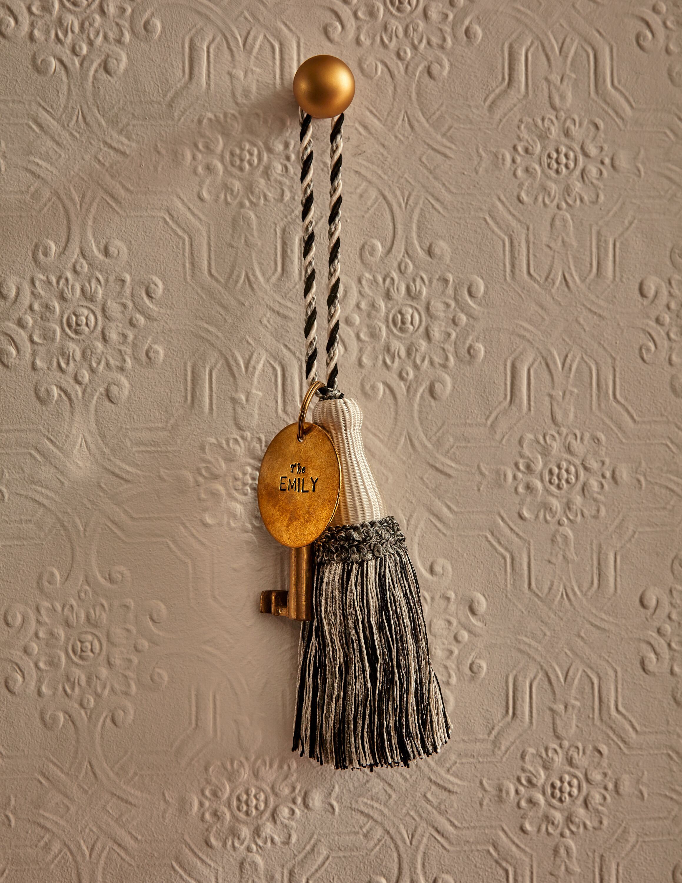 A keychain with a tassel and a round tag with the name "The Emily" on a textured wall.