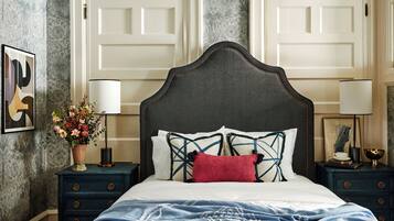 A bedroom with a dark headboard, white bedding, a red pillow, and a blue patterned throw. There are two bedside tables with lamps and a vase of flowers.