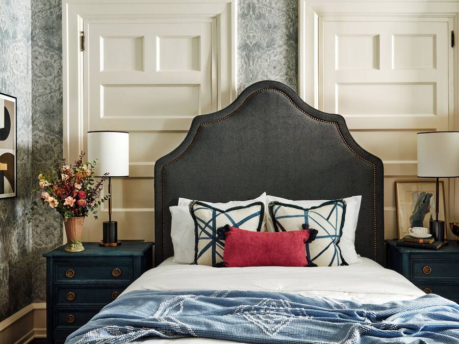 A bedroom with a dark headboard, white bedding, a red pillow, and a blue patterned throw. There are two bedside tables with lamps and a vase of flowers.