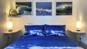 Un dormitorio con una cama que presenta un diseño de ola marina azul, dos lámparas de noche y tres cuadros con motivos de olas marinas en la pared.