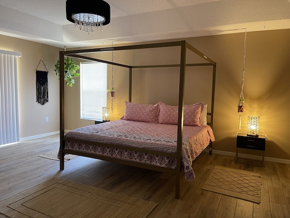 Un letto a baldacchino con copriletto decorato, due comodini con lampade, una pianta pensile in macramè e un pavimento in legno.