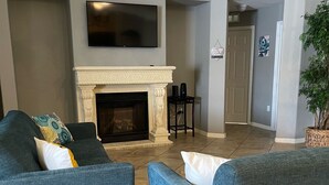 A spacious living room with two blue armchairs, a white fireplace, a flat-screen TV, and a chandelier.