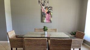 A dining area with a glass-top table surrounded by wicker chairs, a potted plant on the table, and a wall-mounted painting above.