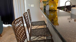 A kitchen with a bar area, wicker chairs, a countertop with a bag of chips, and a window with curtains.