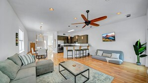 A spacious living area with a sofa, coffee table, and a dining table with chairs. A kitchen with a bar area is visible in the background.