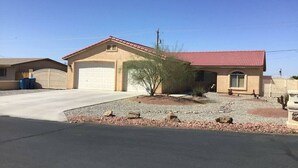 Une maison de plain-pied avec un toit recouvert de tuiles rouges, un garage et un petit jardin agrémenté de rochers et d’un cactus.