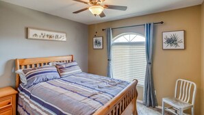 Une chambre à coucher avec un lit en bois, une table de chevet, une chaise, un ventilateur de plafond et deux cadres photos fixés au mur.