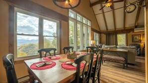 A dining area with a wooden table set for four, large windows offering a view of the landscape, and a cozy seating area with a sofa and armchairs.