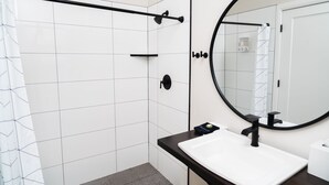 Un baño moderno con ducha, un lavamanos blanco, un grifo negro y un espejo grande.