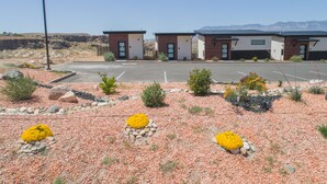 Un parking avec trois bâtiments, une zone de gravier parsemée de rochers et de fleurs jaunes, et des montagnes en arrière-plan.