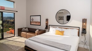 Una habitación de hotel moderna con una cama grande, un sofá, un balcón con un letrero de salida, un cuadro enmarcado, un espejo redondo y un teléfono en la pared.