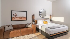 Un dormitorio moderno con una cama, un sofá de cuero marrón, un cuadro enmarcado con un paisaje desértico, un espejo redondo y una mesita de noche con una lámpara.