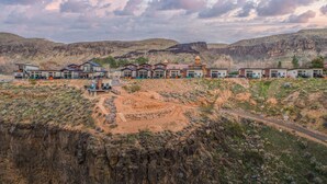Fila de edificios modernos con un sendero de tierra que conduce a un área central, al pie de montañas.
