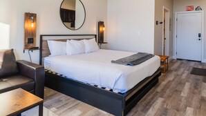 Una habitación de hotel moderna con una cama grande, cabecera de madera, un espejo redondo, luces fijadas a la pared, una mesita pequeña y un letrero de salida.