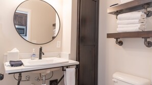 Un baño con un lavabo blanco, un espejo redondo, una puerta oscura y un estante con toallas dobladas.