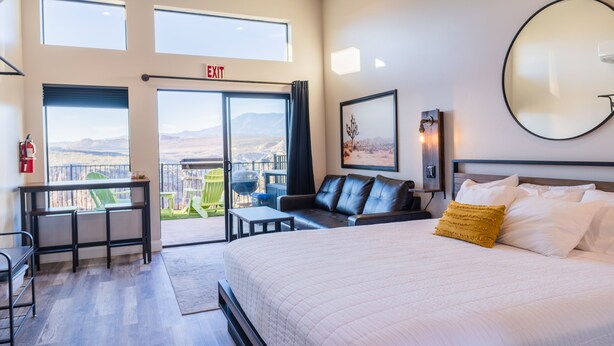 Habitación de hotel con una cama grande, un escritorio, una silla y un balcón con vistas a las montañas.