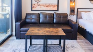 Una habitación de hotel moderna con un sofá de cuero, una mesa de centro de madera, una cama con ropa blanca, un cuadro enmarco de paisaje desértico y un aplique de pared.