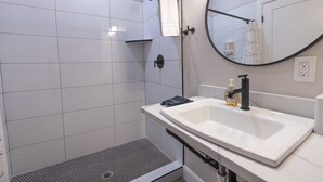 Un baño moderno con un lavamanos blanco, un espejo redondo y una zona de ducha con azulejos blancos.
