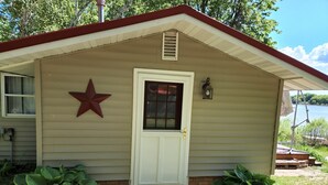 A small, single-story building with a white door, a red star decoration, and a view of a body of water. - Cozy Cabin lake front (Dalbo)