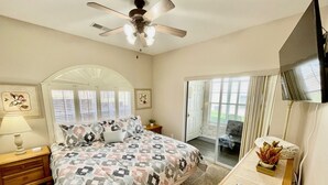 Une chambre à coucher avec un lit, un ventilateur de plafond, une télévision, un bureau et une fenêtre avec des stores.